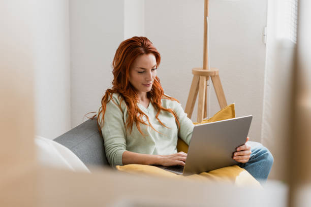 Young woman is working from home, sitting on a couch and typing on a laptop